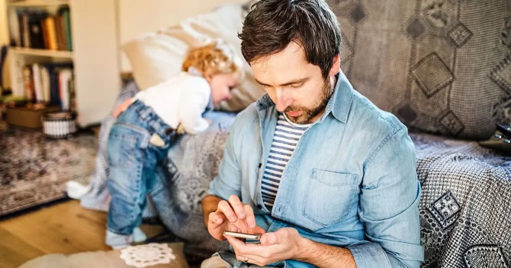 Father looking at his phone while his young son plays beside him, being a present father