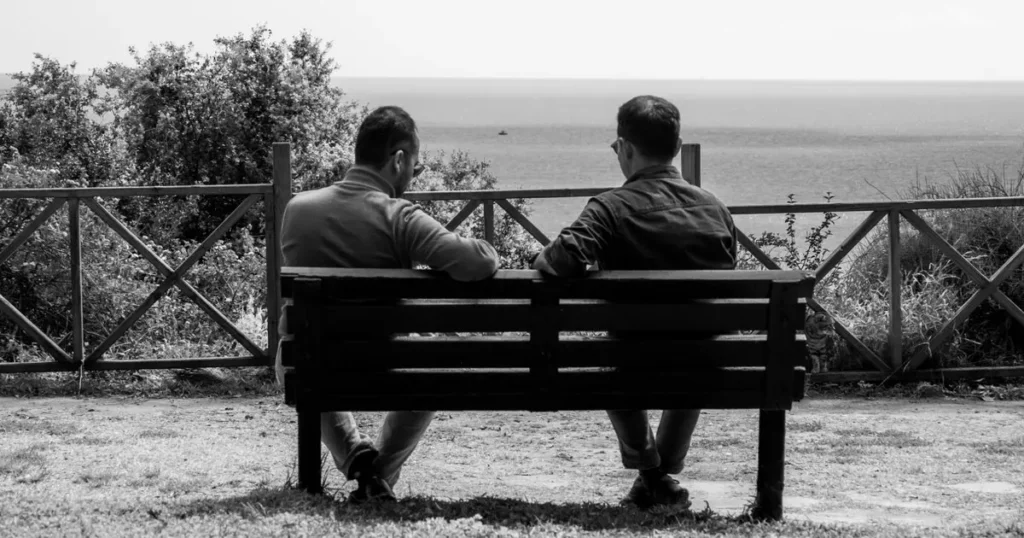 Two male friends talking on a bench overlooking the sea
